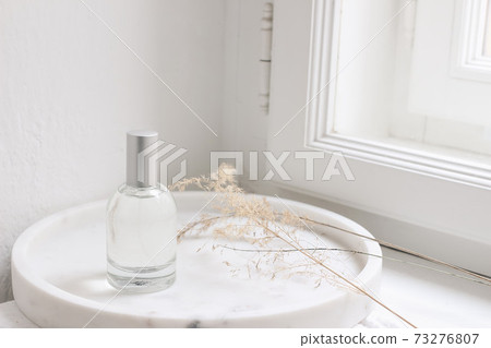 Feminine cosmetic still life scene. Glass bottle, flacon with perfume or eau de toilette and dry grass on white marble tray near window. Selective focus, blurred window sill background. 73276807