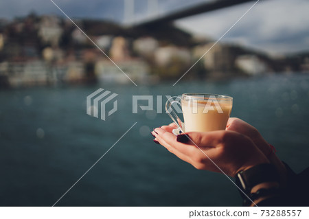 A woman's hand is holding a white cup of hot milky beverage with cinnamon called Turkish salep sahlep on the background of rippling water and misty Maiden s Tower in the distance, Istanbul. A woman's hand is holding a white cup of hot milky beverage with cinnamon called Turkish salep sahlep on the background of rippling water and misty Maiden s Tower in the distance, Istanbul. 73288557