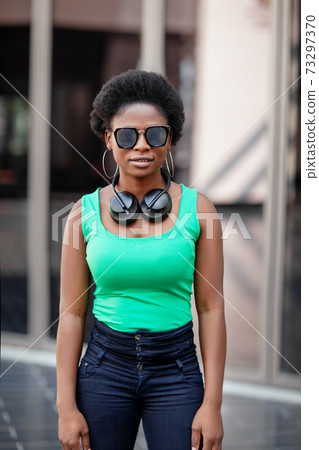 A young African woman wearing headphones and sunglasses smiles happily and looks at the camera 73297370