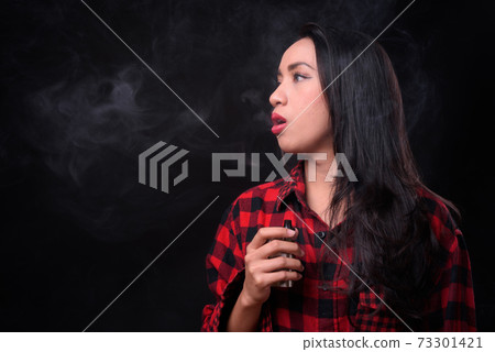Young beautiful Asian woman vaping against black background 73301421