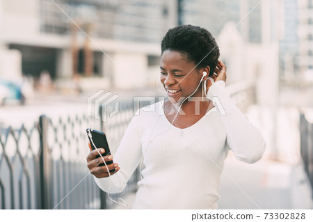 A young African woman in a white sweater and headphones answers a video call while walking down the street. The concept of online communication 73302828