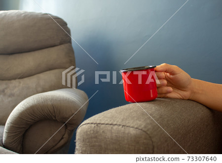 Woman holding a red metal cup resting her hand on the sofa's arm Woman holding a red metal cup resting her hand on the sofa's arm 73307640