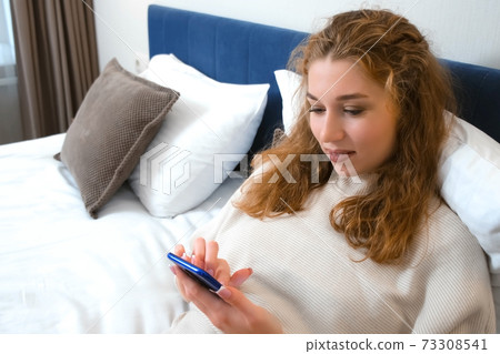 Young smiling woman is browsing smartphone lying in bed in hotel room. 73308541