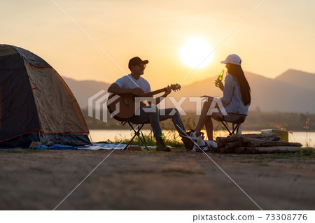 Asian couple with dog playing guitar and drinking beer beside their tent campsite 73308776