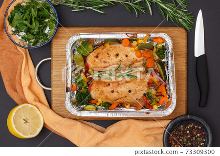 Baked chicken breasts or fillet in metal container with vegetables on cutting board. Baked chicken breasts or fillet in metal container with vegetables on cutting board. 73309300