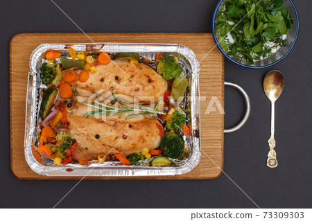 Baked chicken breasts or fillet in metal container with vegetables on cutting board. 73309303