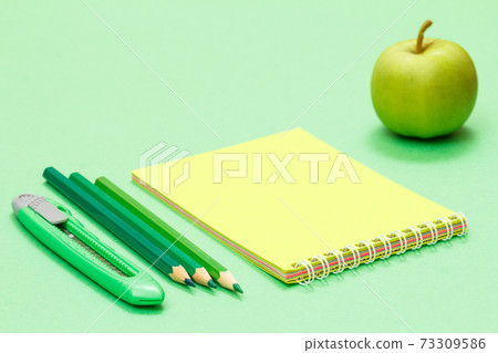 School supplies. Paper knife, color pencils, notebook and apple on the green background. School supplies. Paper knife, color pencils, notebook and apple on the green background. 73309586