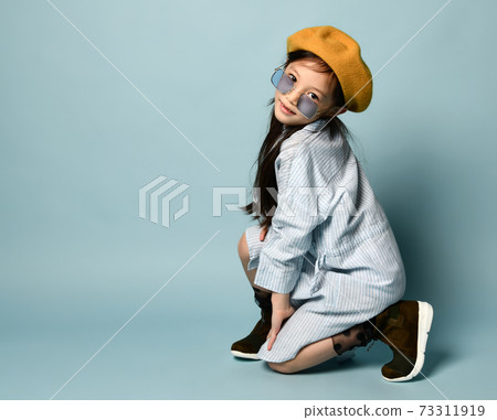Little asian girl in sunglasses, shirt dress, brown beret, boots. Smiling, posing sitting sideways on blue background. Close up Little asian girl in sunglasses, shirt dress, brown beret, boots. Smiling, posing sitting sideways on blue background. Close up 73311919