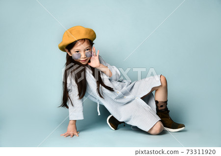 Little asian kid in sunglasses, shirt dress, brown beret, boots. Smiling, posing sitting sideways on blue background. Close up Little asian kid in sunglasses, shirt dress, brown beret, boots. Smiling, posing sitting sideways on blue background. Close up 73311920
