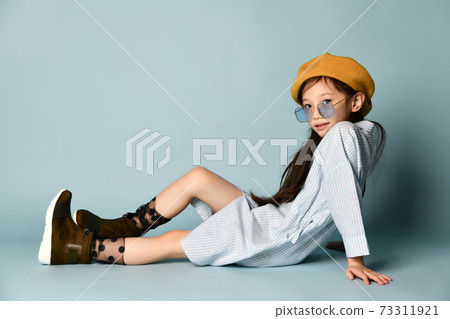 Little asian kid in sunglasses, shirt dress, brown beret, boots. Smiling, posing sitting sideways on blue background. Close up 73311921