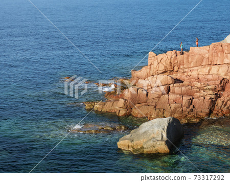 Red Rocks called Rocce Rosse at mediterranean sea coastline in Arbatax with two boys fishing. Tortoli, Ogliastra, Sardinia, Italy. Summer sunny day, golden hour Red Rocks called Rocce Rosse at mediterranean sea coastline in Arbatax with two boys fishing. Tortoli, Ogliastra, Sardinia, Italy. Summer sunny day, golden hour 73317292