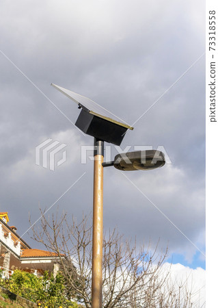 Street lamp with photovoltaic panel close-up for receiving solar energy against the background of a cloudy sky and a cottage on a hill Street lamp with photovoltaic panel close-up for receiving solar energy against the background of a cloudy sky and a cottage on a hill 73318558