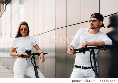 A girl and a guy are walking on electric scooters around the city, a couple in love on scooters. A girl and a guy are walking on electric scooters around the city, a couple in love on scooters. 73319287