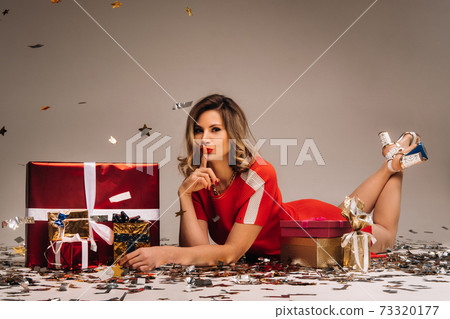 A girl in a red dress is lying on the floor with a bunch of gifts on a gray background in confetti A girl in a red dress is lying on the floor with a bunch of gifts on a gray background in confetti 73320177