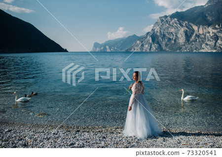 a girl in a smart white dress walks along the embankment of lake Garda.A woman is photographed against the background of a mountain and lake in Italy.Torbole a girl in a smart white dress walks along the embankment of lake Garda.A woman is photographed against the background of a mountain and lake in Italy.Torbole 73320541