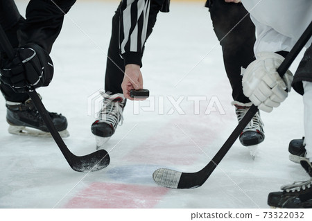 Hand of referee holding puck over ice rink and two hockey players with sticks 73322032