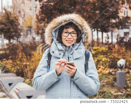 Smiling woman in light blue warm jacket is texting someone on smartphone. Online communication via LTE or Wi-fi. Wireless cellular technology. Autumn season in town. Smiling woman in light blue warm jacket is texting someone on smartphone. Online communication via LTE or Wi-fi. Wireless cellular technology. Autumn season in town. 73322425
