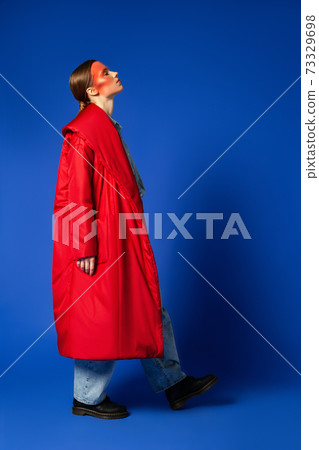 Female model in baggy coat walking against blue background 73329698