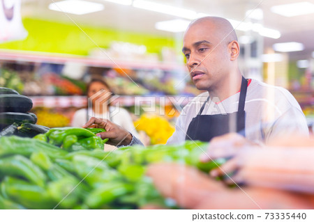 Merchandiser in an apron lays out ripe bell peppers on supermarket counter Merchandiser in an apron lays out ripe bell peppers on supermarket counter 73335440