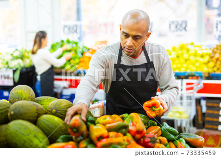 Merchandiser in an apron lays out ripe bell peppers on supermarket counter Merchandiser in an apron lays out ripe bell peppers on supermarket counter 73335459