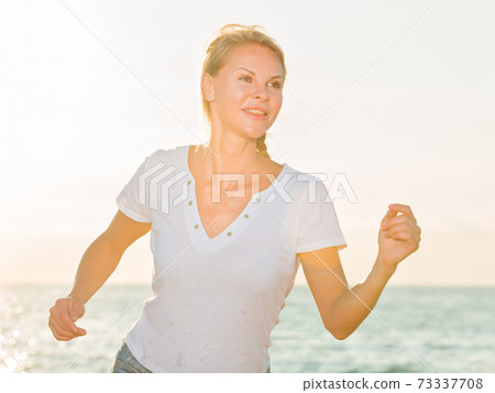 Smiling adult woman in white T-shirt is jogging on the beach Smiling adult woman in white T-shirt is jogging on the beach 73337708