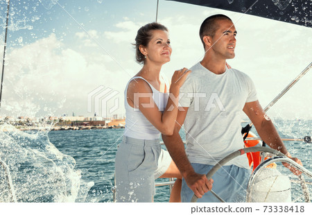 Couple standing at yacht wheel along coast of Barcelona Couple standing at yacht wheel along coast of Barcelona 73338418