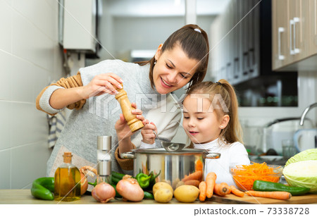 Daughter helping cooking soup and mother add pepper to pan Daughter helping cooking soup and mother add pepper to pan 73338728