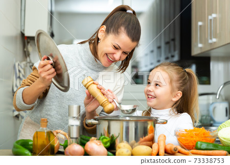 Mother add pepper to vegetable soup and small daughter tasting Mother add pepper to vegetable soup and small daughter tasting 73338753