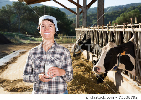 Mature woman farmer with cow milk in glass at cowshed 73338792