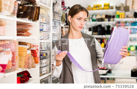 Interested young woman choosing colorful ribbons and braid for dressmaking in sewing supplies shop Interested young woman choosing colorful ribbons and braid for dressmaking in sewing supplies shop 73339528