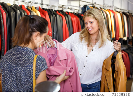 Smiling shopping assistant demonstrating female customer leather jackets in store Smiling shopping assistant demonstrating female customer leather jackets in store 73339558