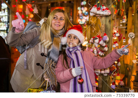 Happy daughter with mom point finger at selected christmas tree decorations at street market Happy daughter with mom point finger at selected christmas tree decorations at street market 73339770