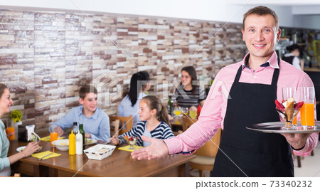 Enthusiastic waiter showing hospitality and meeting visitors in cafe Enthusiastic waiter showing hospitality and meeting visitors in cafe 73340232