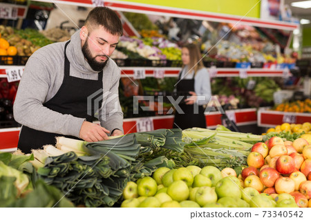 Male shop assistant in apron lays fresh leek in supermarket Male shop assistant in apron lays fresh leek in supermarket 73340542
