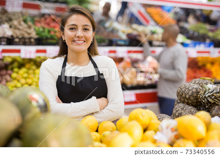 Supermarket woman worker in black apron with melons and lemons in his department Supermarket woman worker in black apron with melons and lemons in his department 73340556