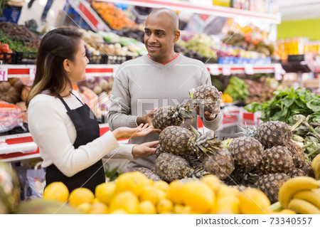 Female grocery salesperson advises man to buy pineapple Female grocery salesperson advises man to buy pineapple 73340557