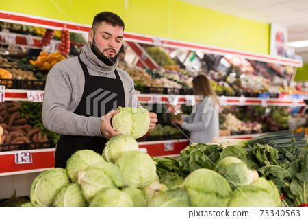Male shop assistant lays fresh cabbage on counter in grocery shop Male shop assistant lays fresh cabbage on counter in grocery shop 73340563