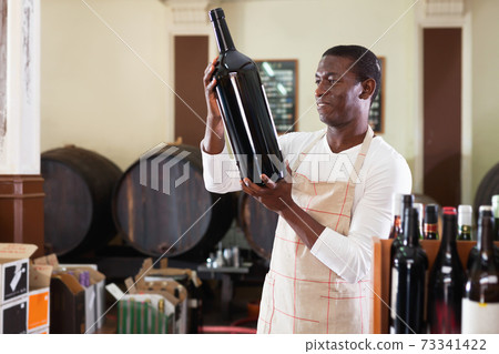 Smiling salesman in apron proposing wine in bottles in winery shop Smiling salesman in apron proposing wine in bottles in winery shop 73341422