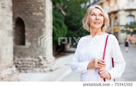 Cheerful mature female is walking in white blouse on the street outdoor Cheerful mature female is walking in white blouse on the street outdoor 73341502