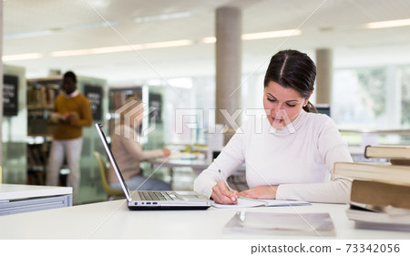 Adult female student with laptop and books in public library 73342056