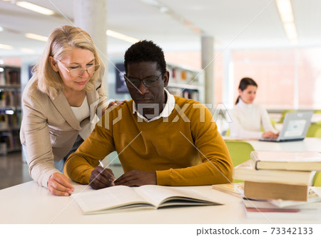 Female teacher working with african-american man in university library Female teacher working with african-american man in university library 73342133