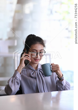Portrait of businesswoman in eye glasses talking on mobile phone and drinking coffee while sitting in comfortable office. 73344256