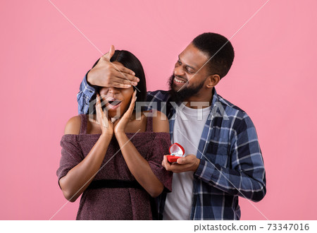 Black guy covering his sweetheart's eyes, giving her engagement ring, making marriage proposal on pink background 73347016