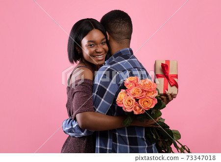 Passionate African American lovers with gift box and beautiful roses for Valentine's Day hugging on pink background 73347018