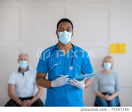 Young African American doctor in antiviral mask and uniform holding clipboard at population immunization centre 73347249