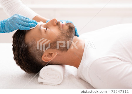 Cosmetologist cleansing bearded man face with toner before treatment Cosmetologist cleansing bearded man face with toner before treatment 73347511