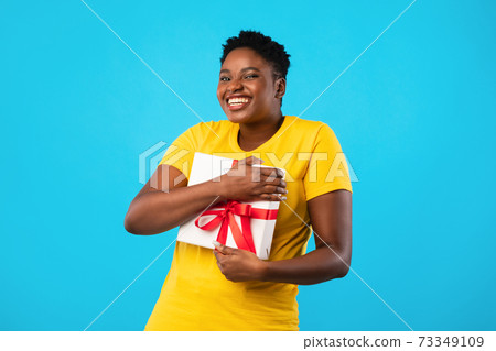 Joyful Plus-Size Black Woman Hugging Gift Box Over Blue Background 73349109