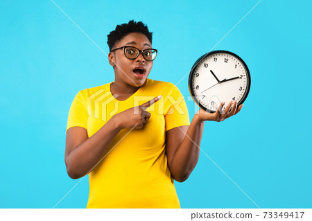 Oversized African Woman Showing Clock Posing Standing Over Blue Background Oversized African Woman Showing Clock Posing Standing Over Blue Background 73349417