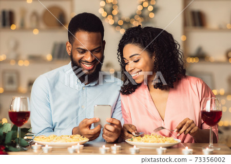 Smiling black man and woman using mobile phone during dinner Smiling black man and woman using mobile phone during dinner 73356002