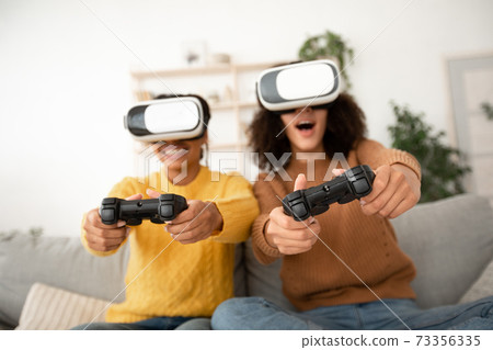 Amazed millennial funny african american sisters with joysticks in vr glasses play in online game 73356335